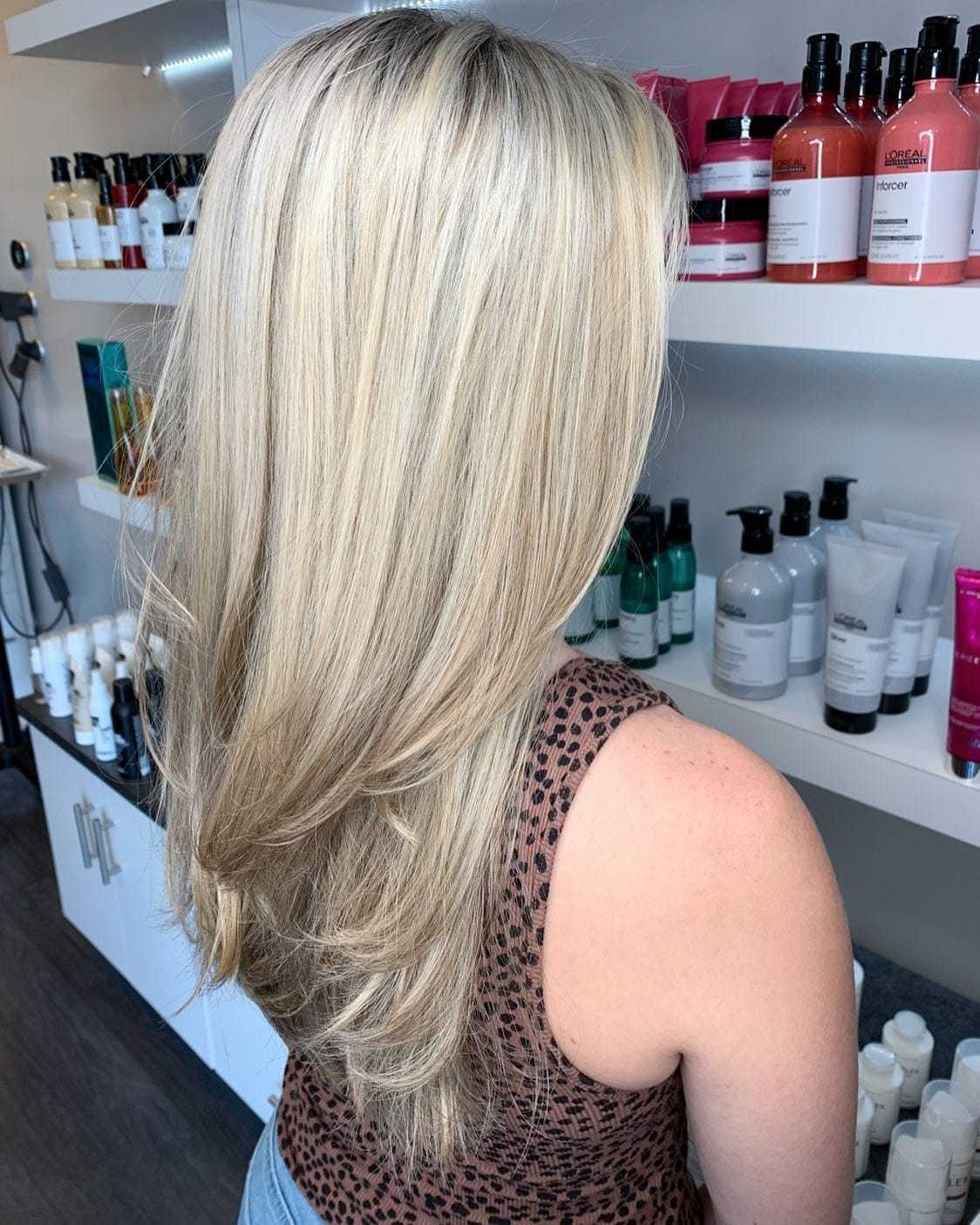Blonde woman with layered hair in a salon, surrounded by hair care products on shelves.