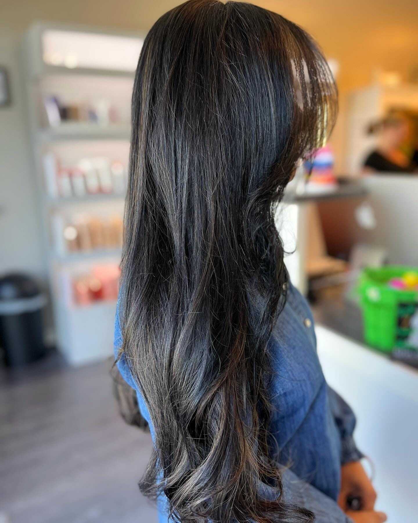 Long, dark, wavy hair styled at a salon. Woman in denim shirt, blurred shelves behind.
