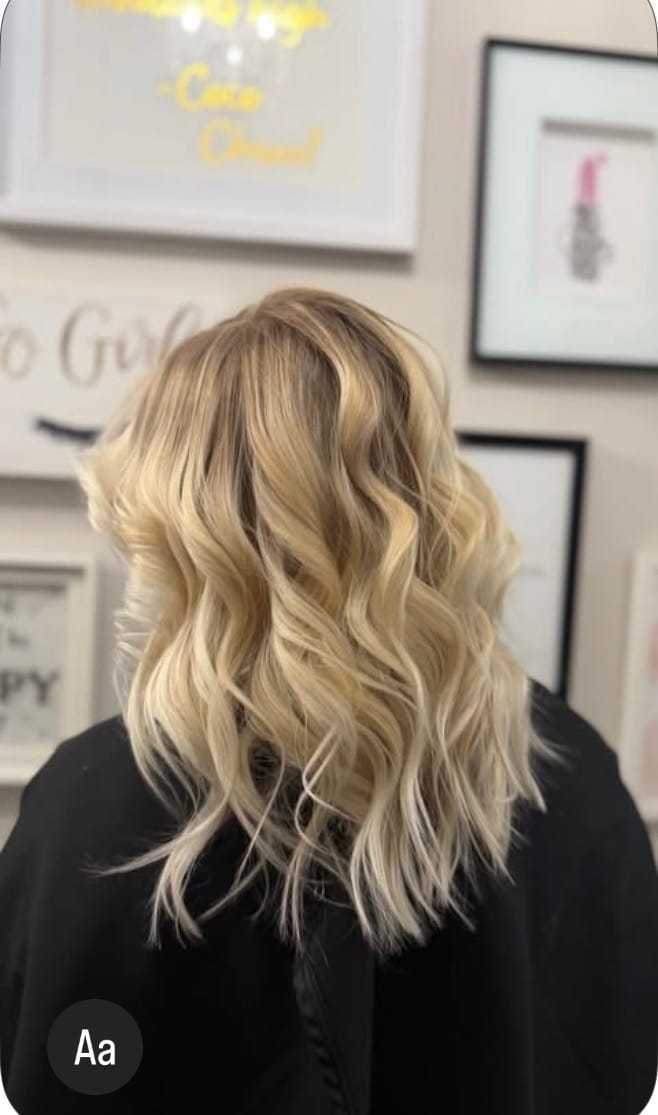 Wavy blonde hairstyle on a person in a salon setting with framed art on the wall.