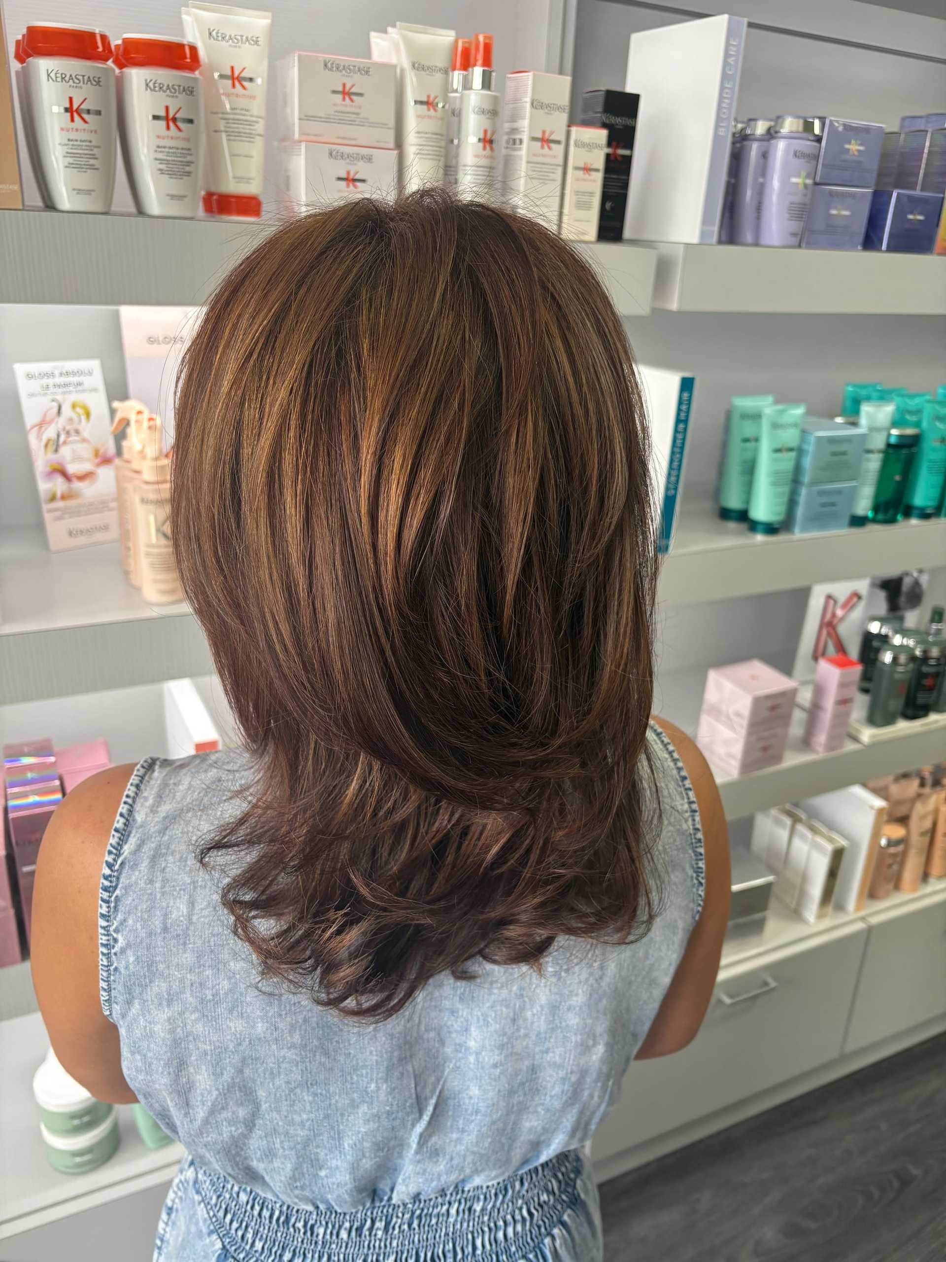 Back view of woman with styled brown hair in a salon, standing before hair care product shelves.
